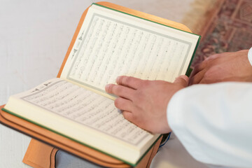 Obraz premium Młody arabski muzułmanin w tradycyjnym stroju czytający świętą księgę Koranu na dywanie modlitewnym przed kolacją iftar podczas uczty ramadanowej w domu