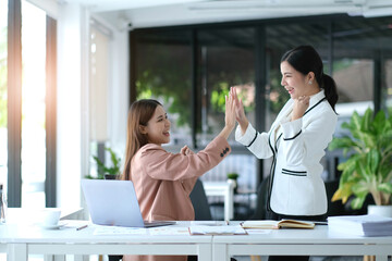 Two young Asian businesswomen show joyful expression of success at work smiling happily with a laptop computer in a modern office.