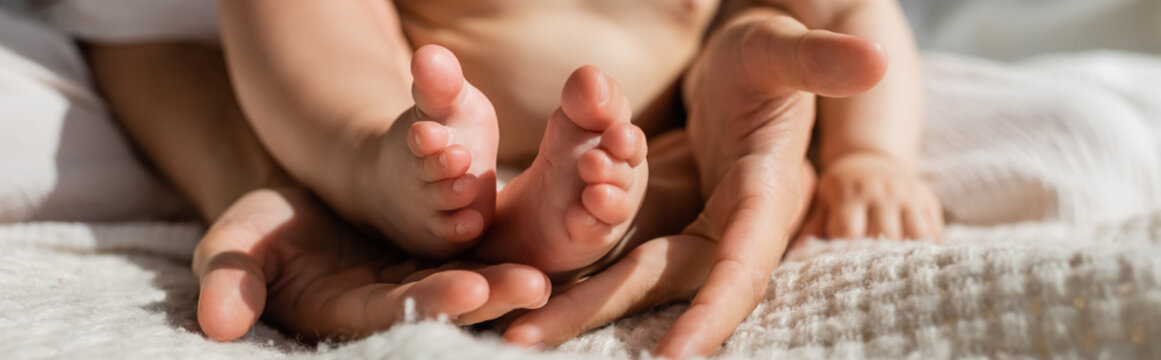 cropped view of mother holding in hands tiny bare feet of infant daughter in bedroom, banner.