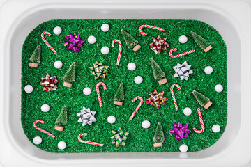 Christmas sensory box. Green colored rice with christmas decorations and toys in sensory bin. Holiday activity for Kids. Sensory play for toddlers. - obrazy, fototapety, plakaty