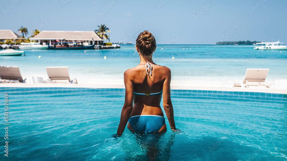 Wall mural Travel vacation - Beautiful young pretty blonde girl back in bikini on her perfect sport sexy body relax in pool near paradise beach at tropical caribbean maldives beach at sunny day in hotel