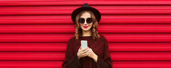 Portrait of stylish smiling woman with smartphone wearing brown knitted poncho, black round hat on...