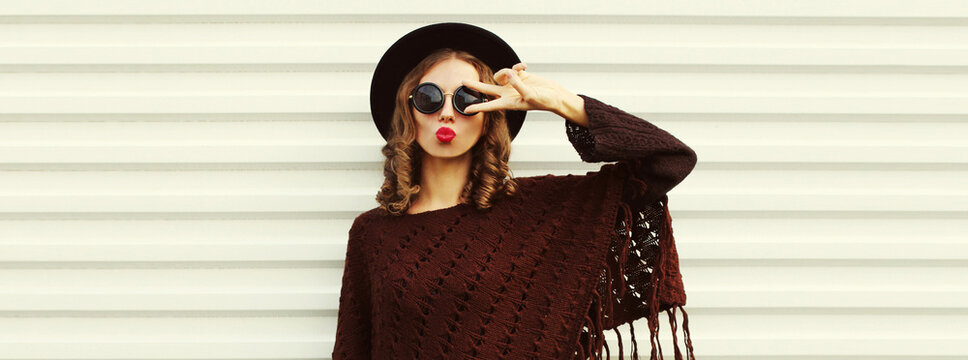 Portrait of beautiful young woman blowing her lips sending sweet air kiss wearing brown knitted poncho, black round hat on white background