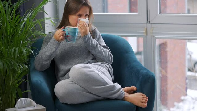 Woman drinks hot tea and coughs into a napkin because she has a cold, flu