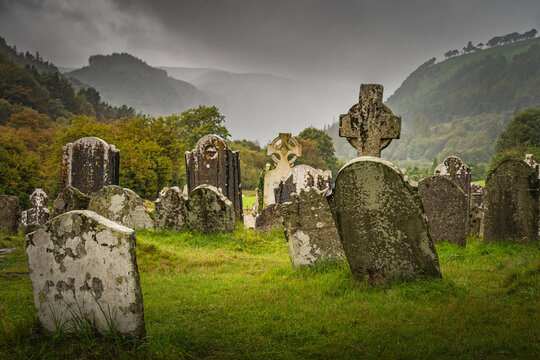 Ancient graves with Celtic crosses in Glendalough Cemetery. Autumn forest, mountains, rain and dramatic storm sky in background, Wicklow, Ireland