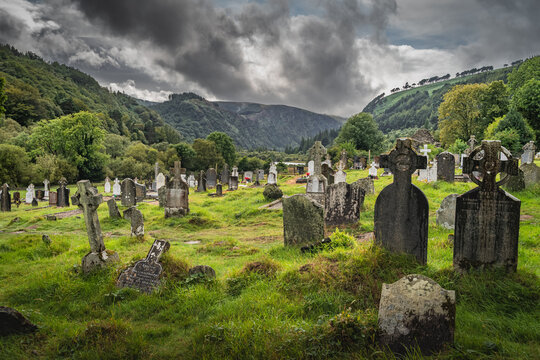 Ancient graves with Celtic crosses in Glendalough Cemetery with mountains and dramatic storm sky in background, Wicklow, Ireland