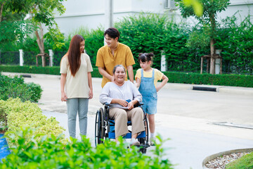 Asian elderly woman sitting on wheelchair which strolling by her happy family when walking at green park.