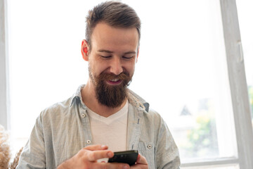 Handsome young bearded man spending time at home with phone in hands.