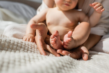 partial view of mother holding in hands tiny bare feet of infant daughter in bedroom.