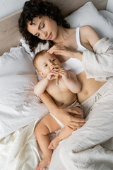 Top view of brunette mom in pajamas touching baby daughter with toy on bed.