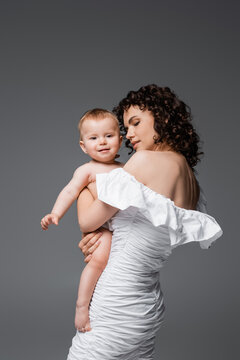 Brunette woman in dress holding smiling infant daughter isolated on grey.