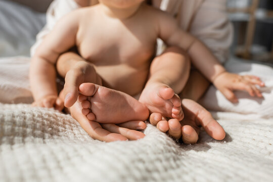 cropped view of mother holding in hands tiny bare feet of infant daughter in bedroom.