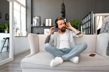 Positive bearded man listening music with headphones while sitting on couch at home