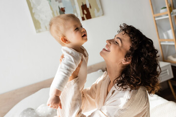 joyful and curly woman in loungewear holding in arms baby daughter in romper.