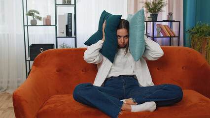 Repair work at neighbours concept. Irritated woman sit on couch cover ears with pillows annoyed by...