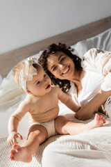 Obraz na płótnie Canvas cheerful mother with curly hair lying on bed near cute baby daughter in headband.