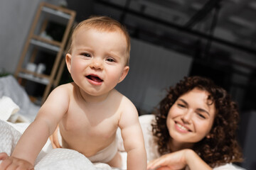 baby girl crawling on bed and looking at camera near blurred mother with curly hair.