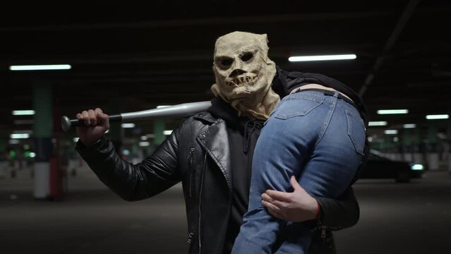 Homicidal maniac on Halloween or Doomsday. Male killer in scary scarecrow mask with baseball bat in his hands carries girl on victim's shoulder in an underground parking lot at night.