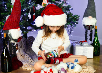 A little girl in a red Santa's Hat is sitting under the Christmas tree and playing with a magic snow globe. Christmas and New Year atmosphere. Concept of a happy childhood. Slow motion