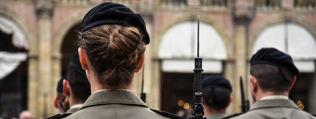 Horizontal banner or header with uniformed women standing during the military ceremony in Bologna, Italy. In the foreground, a woman seen from behind with a bayonet rifle.