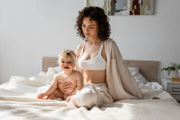 curly mother in loungewear with crop top sitting on bed with infant daughter in headband.