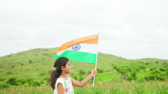 Side view tracking shot of happy running girl kid with indian flag near green fields and mountain - concept of patriotism, independence day celebration and liberty