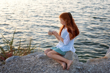 Fototapeta na wymiar A beautiful girl with long red hair sits on the shore of the sea, ocean, river and holds a large shell in her hands. Concept of tourism, travel, vacation by the ocean in summer.
