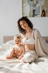 Fototapeta na wymiar curly woman in loungewear with crop top sitting on bed with infant daughter in headband.