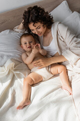 Fototapeta na wymiar high angle view of pleased curly mother in loungewear lying with baby girl on bed.