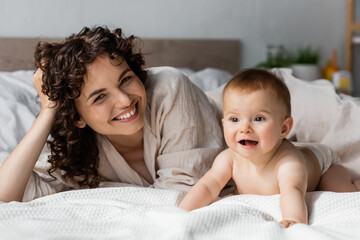 happy woman with curly hair smiling while looking at camera near excited infant baby crawling on bed.