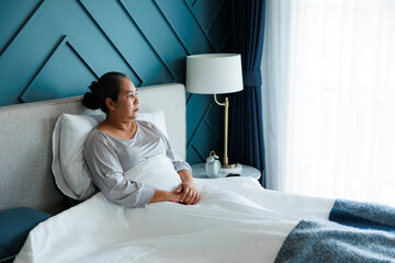 Alone retired asian elder woman depressed looking out to window miss daughter feel sadness, sitting on bed at home.