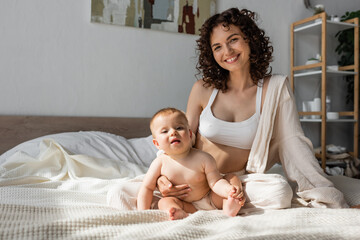 Obraz na płótnie Canvas positive mother in loungewear with crop top sitting near infant daughter in bedroom.