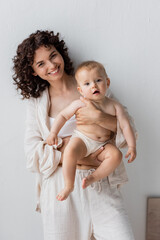 Smiling woman in pajamas holding baby girl and looking at camera at home.