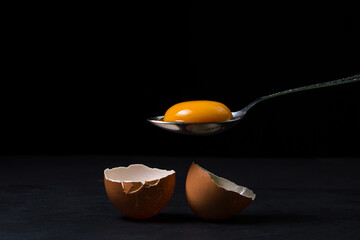 Egg yolk in a spoon next to the shell on a black background. Broken chicken egg. - obrazy, fototapety, plakaty