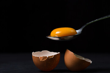 Egg yolk in a spoon next to the shell on a black background. Broken chicken egg.