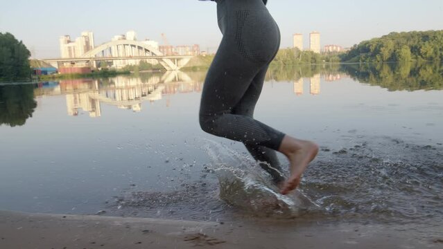 unrecognizable young woman in athletic gray tight leggings runs through water on sandy beach by river. Close up girl's legs run and she goes in for sports in morning outdoors in summer.