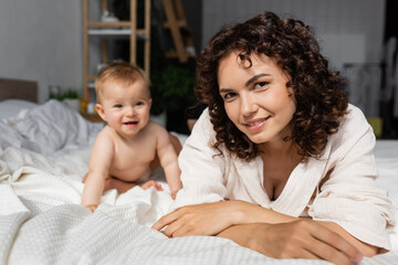 cheerful mother with curly hair looking at camera joyful baby daughter on bed.