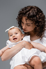 Curly parent looking at baby girl in white headband and dress isolated on grey.