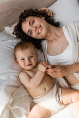 Obraz na płótnie Canvas top view of pleased curly mother in loungewear lying with baby girl on bed.