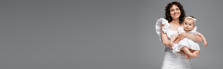 Curly mom in white dress holding baby girl isolated on grey, banner.