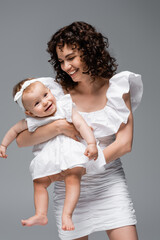 Smiling mother looking at cute baby girl in dress isolated on grey.