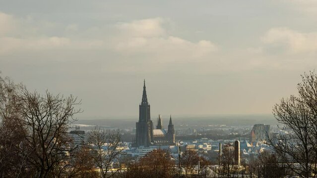 Time lapse shot of Ulm minster in city center seen from Kuhberg through some trees on a golden morning in winter with clouds and some sun rays