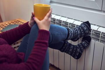 Woman heating feet on a chilly winter day, holding coffee tea cup, energy and gas crisis, cold room, heating problems.