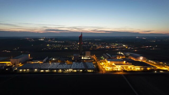 Aerial time lapse shot of an industrial zone with a big silo tower at night. Busy streets and illuminated villages are in the background