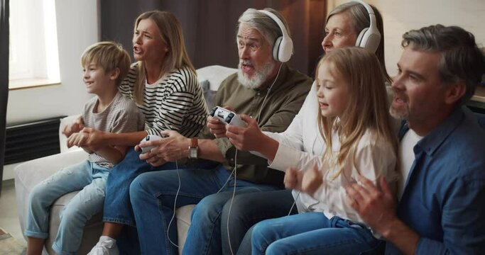 Mature man and woman in headphones playing video games while family cheering them during leisure at home