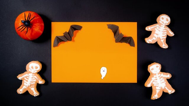 4k White painted ghost on orange background. Black rectangular frame adorned with orange pumpkin with a black spider and skeleton-shaped gingerbread. Halloween holiday concept. Stop motion animation.