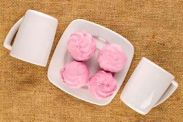 Several sweet marshmallows with white saucer and two cups on jute cloth, macro, top view.