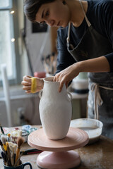 Young woman works with big vase using special tool to make pitcher smooth shape. Brown-haired craftswoman with concerned expression puts big effort into clay vase to send to exhibition in museum
