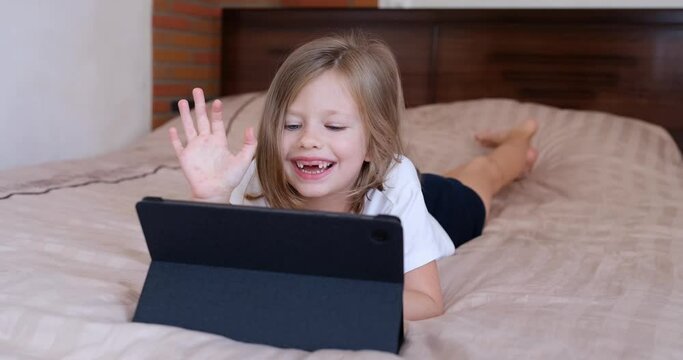 Happy caucasian girl child at home lying on bed making video call using tablet and waving