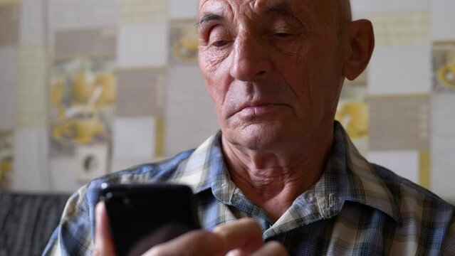 Portrait Of A Caucasian Pensioner 70 Years Old Holding A Smartphone In His Hands Clicking On The Screen While Inside. An Active Elderly Man Uses Social Networks, Reads News Using A Smartphone